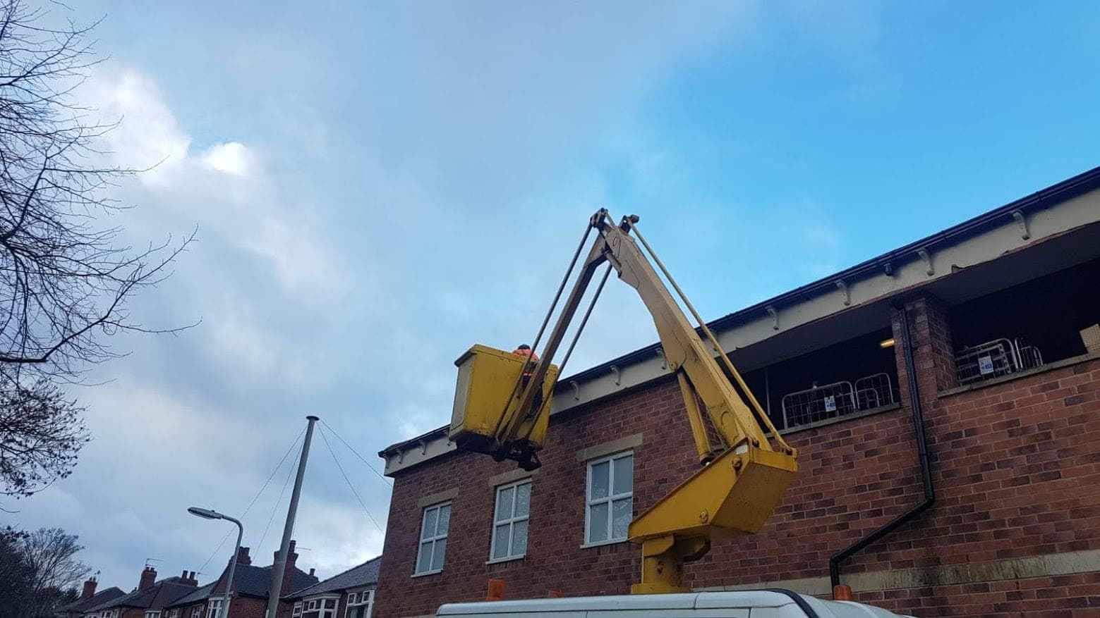 A cherry picker raised to the eaves of a two-storey brick commercial building for powered access to the gutters